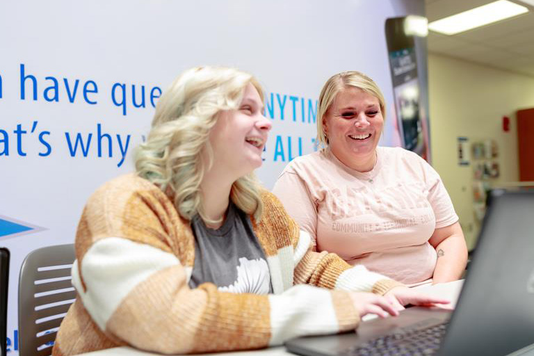 wo smiling people sitting at a table looking at a laptop together.