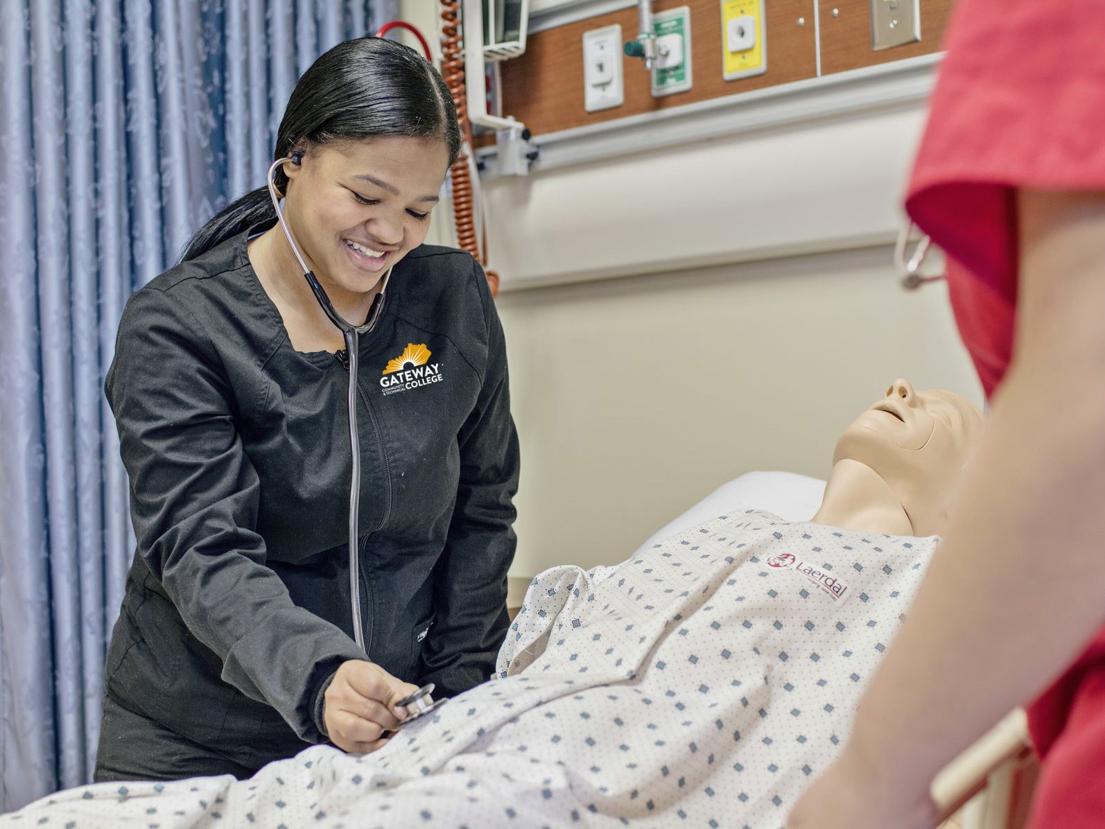 A nursing student from Gateway College practices using a stethoscope on a medical training mannequin in a simulated hospital room. The student is smiling and focused, wearing black scrubs with the Gateway College logo. Another person in red scrubs is partially visible, observing the activity. The mannequin is dressed in a hospital gown marked “Laerdal,” a brand known for medical simulation equipment.