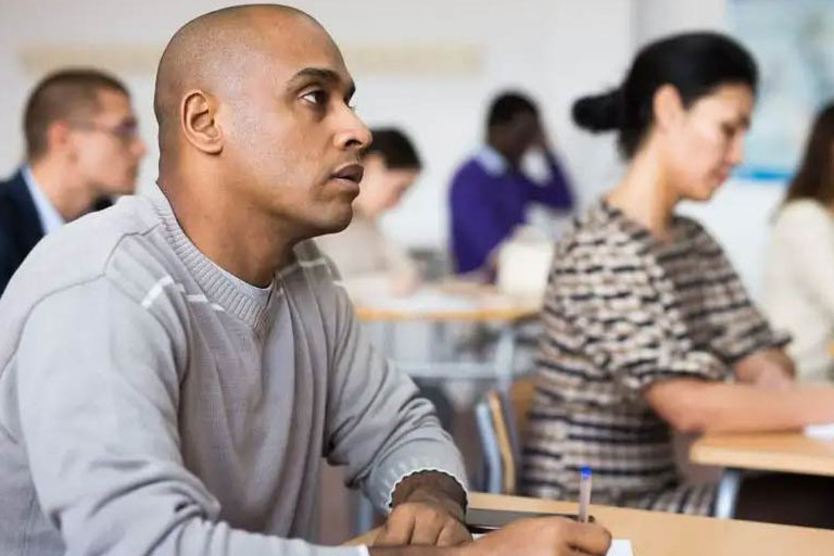 A group of people sitting at desks in a classroom, focused on their work. The foreground features a person in a gray sweater writing on paper, while others in the background are also engaged in similar activities, with a whiteboard visible on the wall.